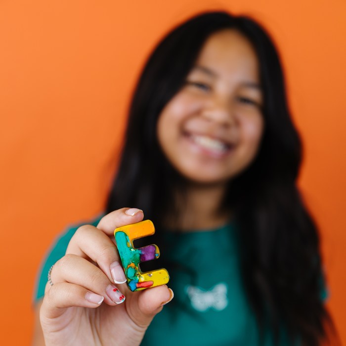 Girl smiling and holding up a single rainbow crayon letter E
